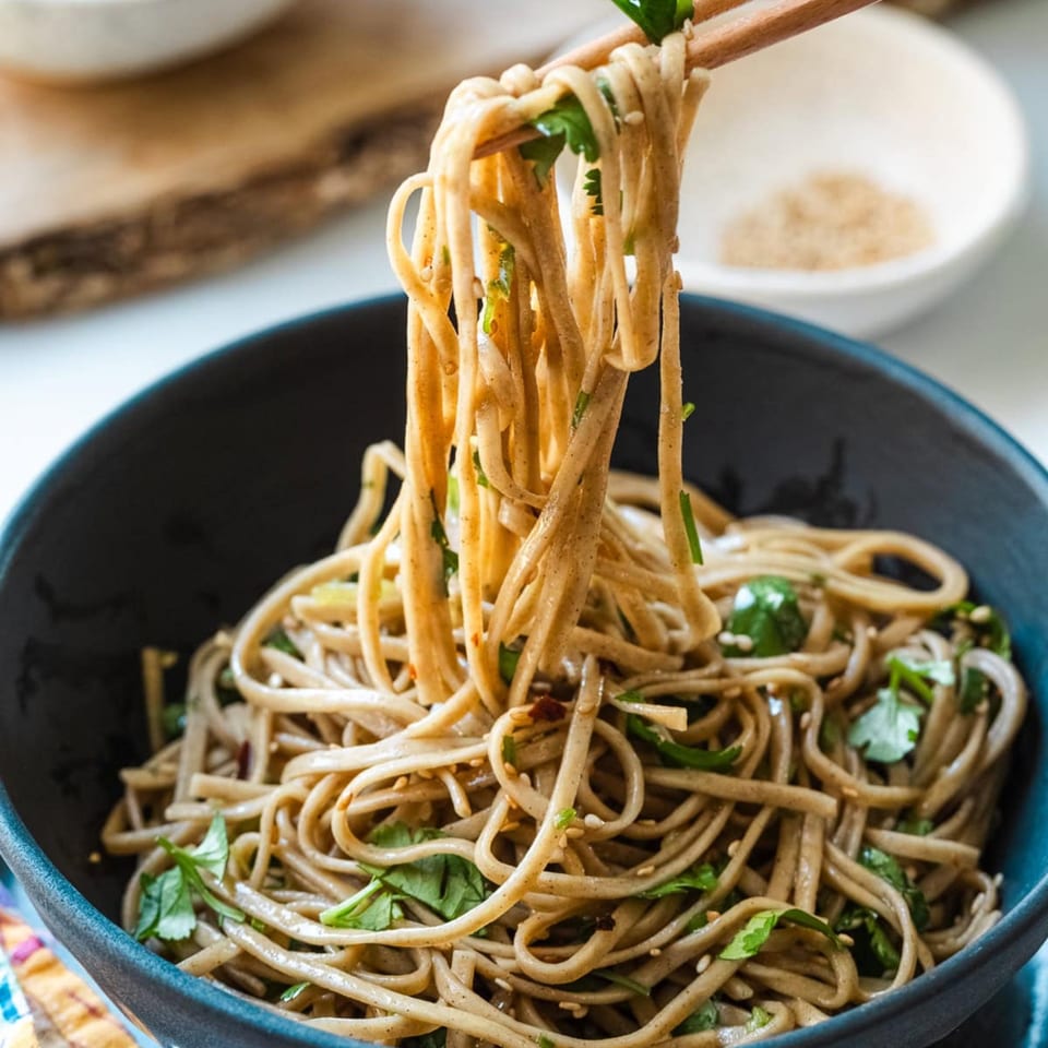 Chopsticks lifting Japanese soba noodles with cilantro and sesame seeds from a dark bowl.