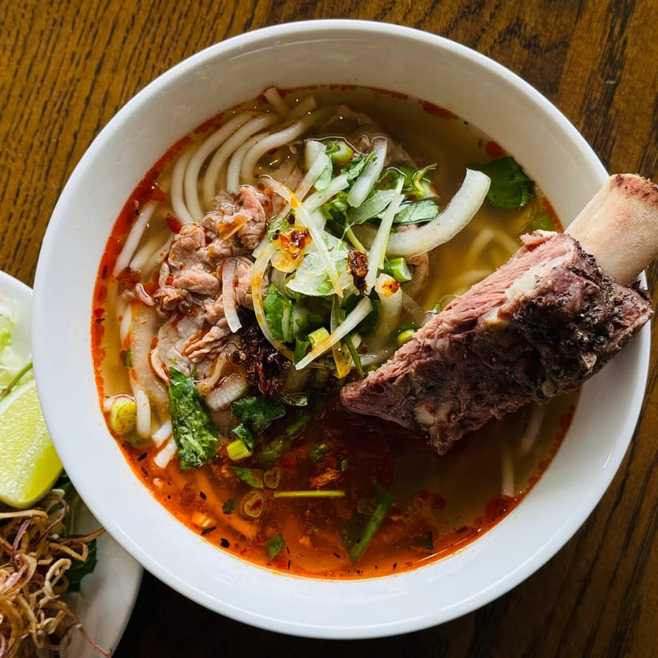 A close-up of a Japanese noodle soup with beef, vegetables, and a large bone in a white bowl.