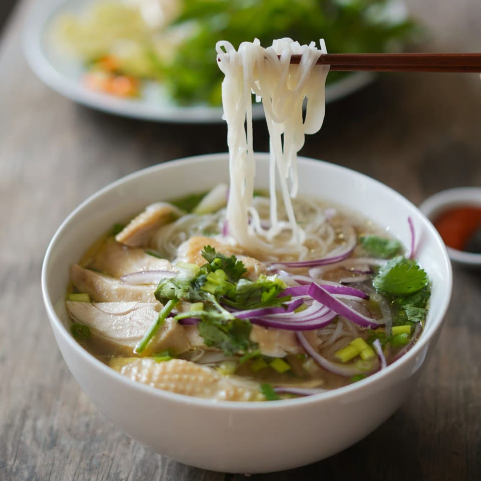 Chopsticks lifting rice noodles from a bowl of chicken pho with red onions and cilantro.