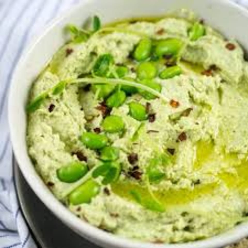 A white bowl of green edamame hummus, drizzled with olive oil and garnished with edamame and herbs.