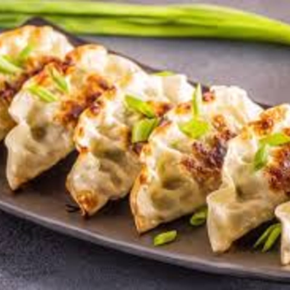 A row of golden-brown Japanese gyoza dumplings garnished with chopped green onions on a dark plate.