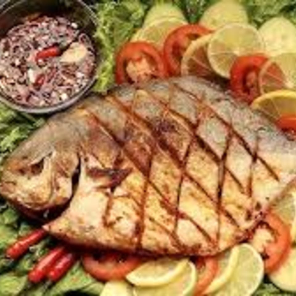 Grilled whole fish with scored skin, served on lettuce with lemon, tomato, and a side of sauce.